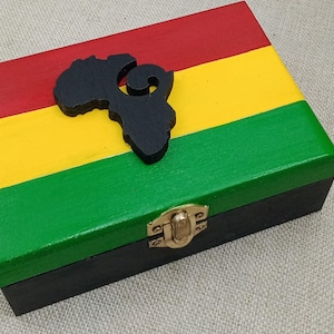 Könnte beinhalten: Eine Holzkiste mit einem schwarz-grün-gelb-roten Streifenmuster. Die Kiste hat eine schwarze Silhouette Afrikas mit einem Wirbelmuster oben. Die Kiste hat einen goldenen Verschluss.