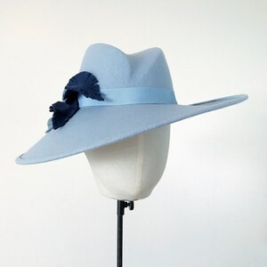Pu&ograve; includere: Un cappello fedora azzurro con tesa larga, fascia in nastro abbinata e abbellimenti a forma di foglia blu scuro. Il cappello &egrave; esposto su una testa di manichino bianca su sfondo bianco.