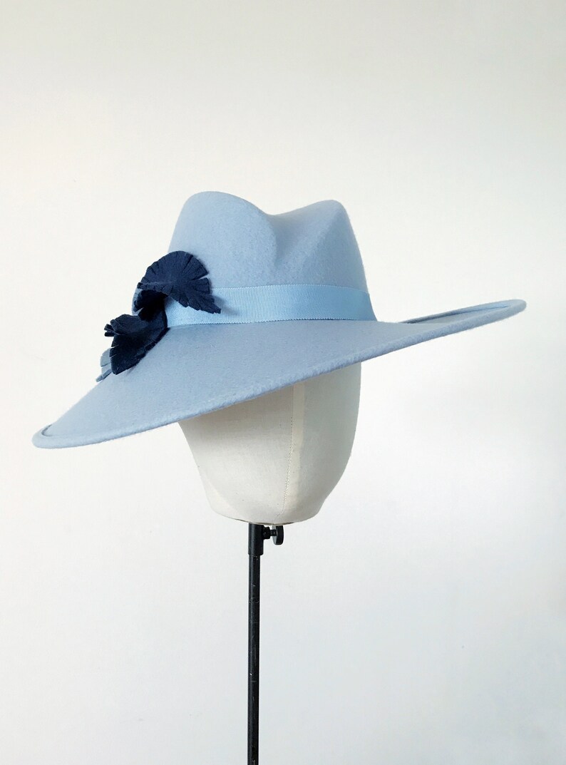 Pu&ograve; includere: Un cappello fedora azzurro con tesa larga, fascia in nastro abbinata e abbellimenti a forma di foglia blu scuro. Il cappello &egrave; esposto su una testa di manichino bianca su sfondo bianco.