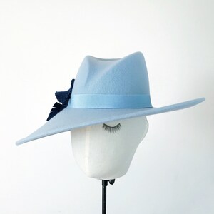 Pu&ograve; includere: Un cappello fedora azzurro con fascia in nastro abbinata e un dettaglio decorativo blu scuro. Il cappello &egrave; esposto su una testa di manichino bianca con gli occhi chiusi, su sfondo bianco. Il cappello ha una tesa ampia e piatta.