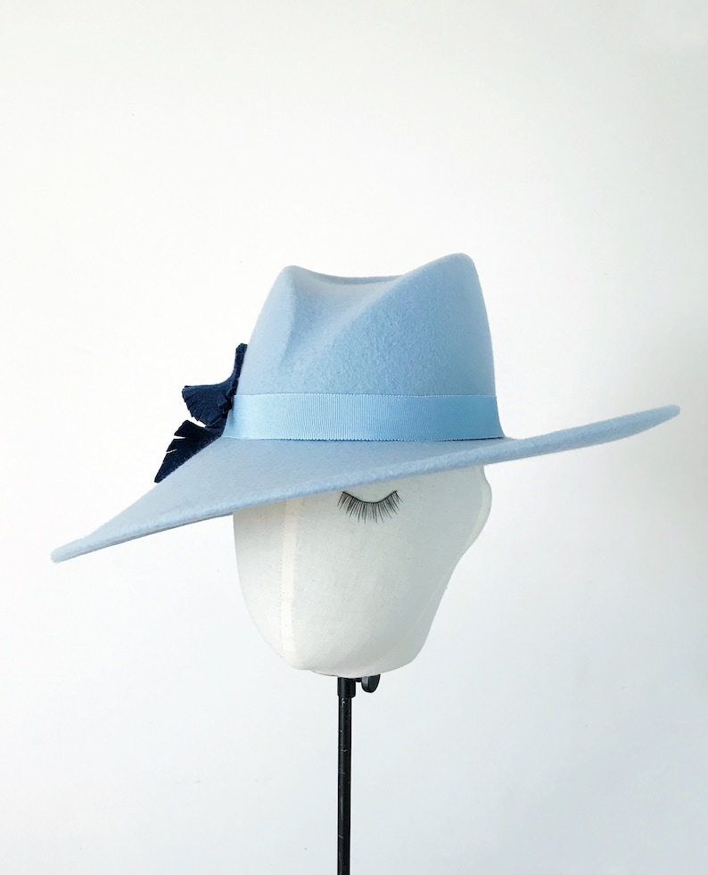Pu&ograve; includere: Un cappello fedora azzurro con fascia in nastro abbinata e un dettaglio decorativo blu scuro. Il cappello &egrave; esposto su una testa di manichino bianca con gli occhi chiusi, su sfondo bianco. Il cappello ha una tesa ampia e piatta.