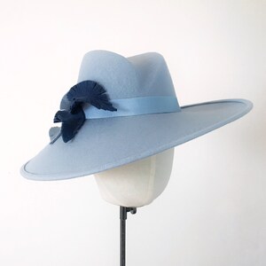 Pu&ograve; includere: Un cappello fedora azzurro con tesa larga, nastro blu scuro e decorazioni a foglie. Il cappello &egrave; esposto su una testa di manichino. Il cappello ha una classica forma fedora ed &egrave; un accessorio elegante.