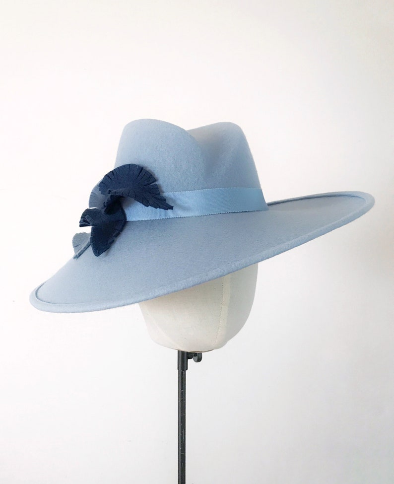 Pu&ograve; includere: Un cappello fedora azzurro con tesa larga, nastro blu scuro e decorazioni a foglie. Il cappello &egrave; esposto su una testa di manichino. Il cappello ha una classica forma fedora ed &egrave; un accessorio elegante.