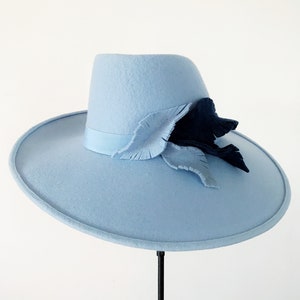 Pu&ograve; includere: Un cappello azzurro con tesa larga e corona strutturata. Il cappello &egrave; adornato da un motivo a foglie decorative blu scuro e azzurro. Il cappello &egrave; esposto su un supporto nero su sfondo bianco.