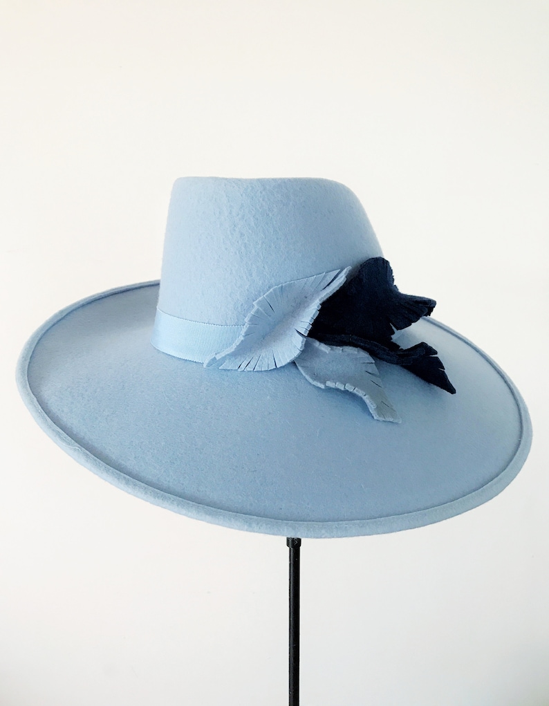 Pu&ograve; includere: Un cappello azzurro con tesa larga e corona strutturata. Il cappello &egrave; adornato da un motivo a foglie decorative blu scuro e azzurro. Il cappello &egrave; esposto su un supporto nero su sfondo bianco.