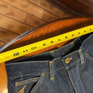 May include: Dark blue denim jeans with a yellow measuring tape showing a measurement of approximately 12 inches. The jeans have a button closure, pockets, and gold stitching. A gold emblem is visible on the back pocket.