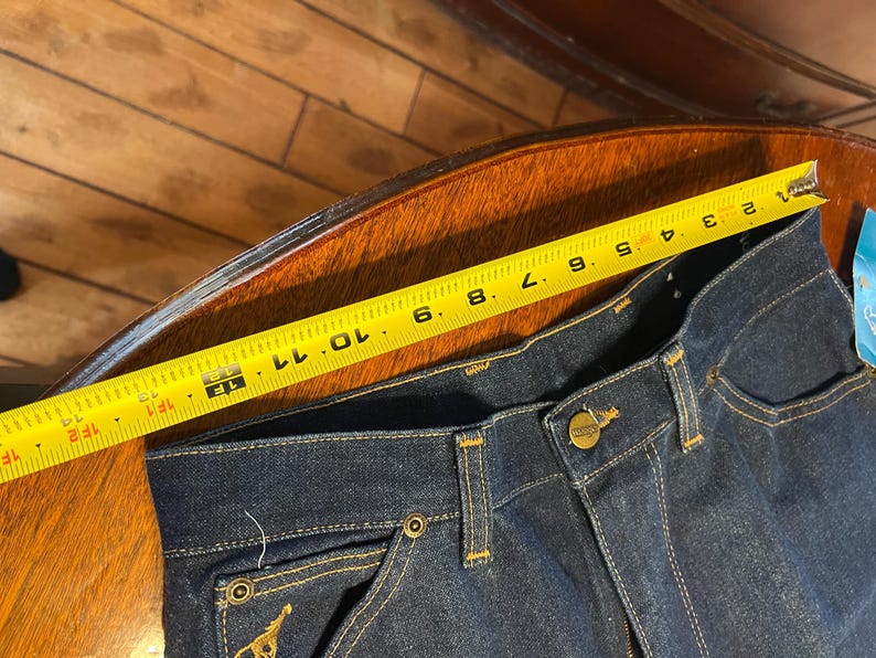 May include: Dark blue denim jeans with a yellow measuring tape showing a measurement of approximately 12 inches. The jeans have a button closure, pockets, and gold stitching. A gold emblem is visible on the back pocket.