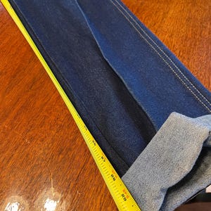 May include: Dark blue denim jeans with a folded cuff. A yellow measuring tape is laid across the jeans, showing measurements in inches. The jeans have white stitching along the seams.
