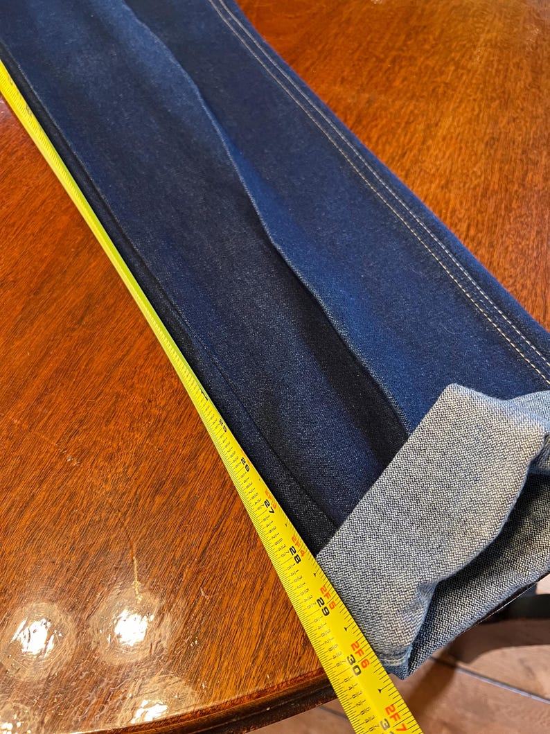 May include: Dark blue denim jeans with a folded cuff. A yellow measuring tape is laid across the jeans, showing measurements in inches. The jeans have white stitching along the seams.