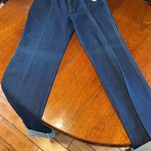 May include: Dark blue Wrangler jeans with a regular fit. The jeans have a classic five-pocket design, a zipper fly, and a Wrangler tag. The cuffs are rolled up, and the jeans are displayed on a wooden table.