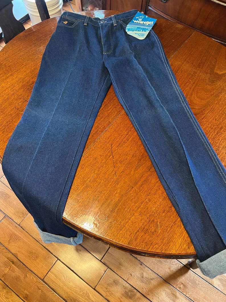 May include: Dark blue Wrangler jeans with a regular fit. The jeans have a classic five-pocket design, a zipper fly, and a Wrangler tag. The cuffs are rolled up, and the jeans are displayed on a wooden table.