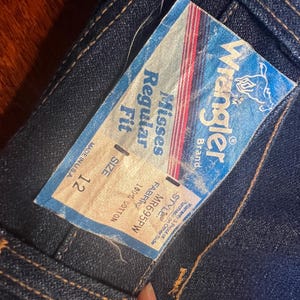 May include: Close-up of a pair of dark blue denim jeans with a Wrangler brand tag. The tag reads "Misses Regular Fit, Size 12, 100% Cotton, Made in USA." The tag has a blue background with red and white stripes.