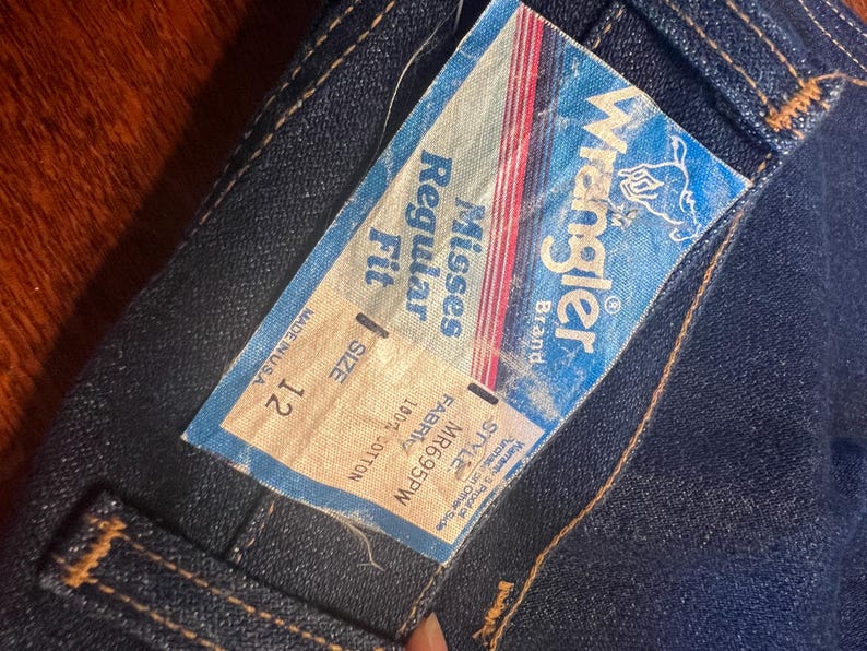 May include: Close-up of a pair of dark blue denim jeans with a Wrangler brand tag. The tag reads "Misses Regular Fit, Size 12, 100% Cotton, Made in USA." The tag has a blue background with red and white stripes.