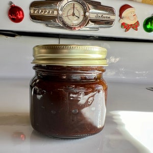May include: A small, clear glass jar filled with a dark brown, viscous substance, likely a preserve or spread. The jar has a gold-colored metal lid and is set against a white background. Christmas ornaments are visible in the background.