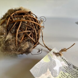 May include: A spherical bird nesting ball made of woven twigs and wool. The ball is brown and tan, with a tag attached by twine. The tag has text that reads "Nesting Wool".