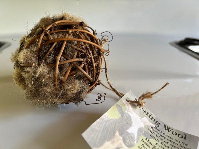 May include: A spherical bird nesting ball made of woven twigs and wool. The ball is brown and tan, with a tag attached by twine. The tag has text that reads "Nesting Wool".