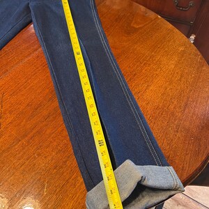 May include: Dark blue denim jeans with a straight leg and white stitching. The jeans are measured with a yellow tape measure, showing a length of approximately 42 inches. The cuffs are folded up.