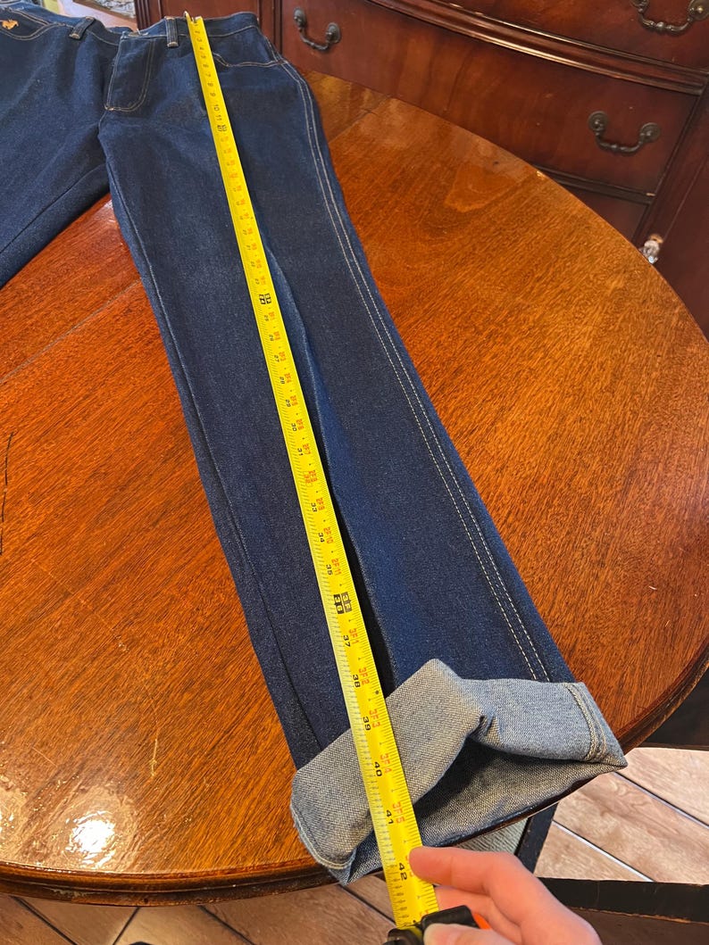 May include: Dark blue denim jeans with a straight leg and white stitching. The jeans are measured with a yellow tape measure, showing a length of approximately 42 inches. The cuffs are folded up.
