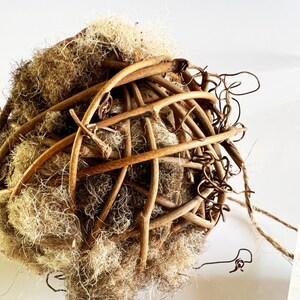 May include: A spherical decorative object composed of interwoven brown twigs and a central mass of light brown, fibrous material. The twigs form a cage-like structure around the core, with some extending outwards. A natural, rustic aesthetic.