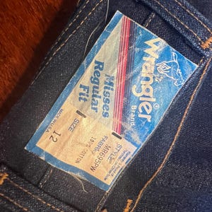 May include: Close-up of dark blue denim jeans with a Wrangler brand tag attached. The tag reads "Misses Regular Fit" and "Size 12". The jeans are made of 100% cotton and are made in the USA.