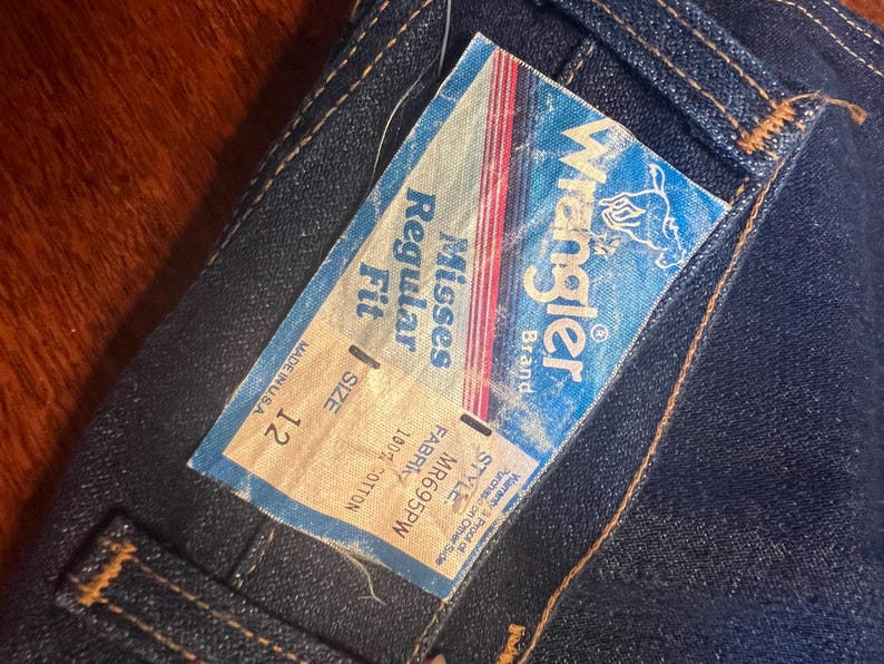 May include: Close-up of dark blue denim jeans with a Wrangler brand tag attached. The tag reads "Misses Regular Fit" and "Size 12". The jeans are made of 100% cotton and are made in the USA.