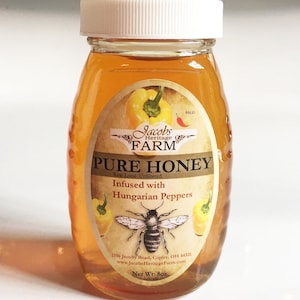 May include: A glass jar of honey infused with Hungarian peppers. The label reads "Jacobs Heritage Farm Pure Honey Raw Local Unfiltered Infused with Hungarian Peppers" and includes a bee illustration. The jar is 8 ounces.