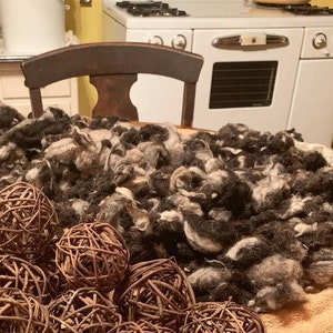May include: A pile of raw wool in shades of black, gray, and white sits on a wooden chair. Wicker spheres are in the foreground. A vintage white stove is in the background.