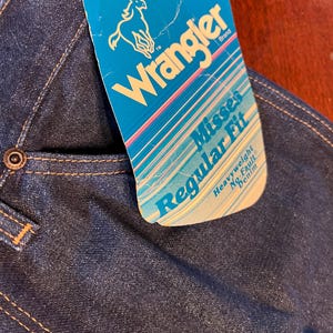 May include: Close-up of dark blue denim jeans with a Wrangler brand tag. The tag is turquoise with the Wrangler logo and text that reads "Misses Regular Fit Heavyweight No-Fault Denim". The jeans have a button closure and visible stitching.