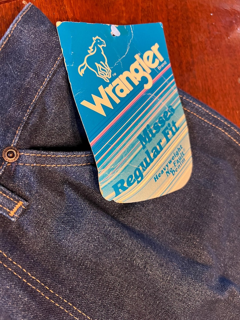May include: Close-up of dark blue denim jeans with a Wrangler brand tag. The tag is turquoise with the Wrangler logo and text that reads "Misses Regular Fit Heavyweight No-Fault Denim". The jeans have a button closure and visible stitching.