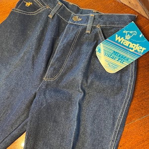May include: Dark blue denim Wrangler jeans with a gold embroidered horse logo on the pocket. The jeans have a classic five-pocket design, a button and zipper closure, and a "Wrangler" brand tag. The jeans are labeled "Misses Regular Fit Heavyweight No-Fault Denim."