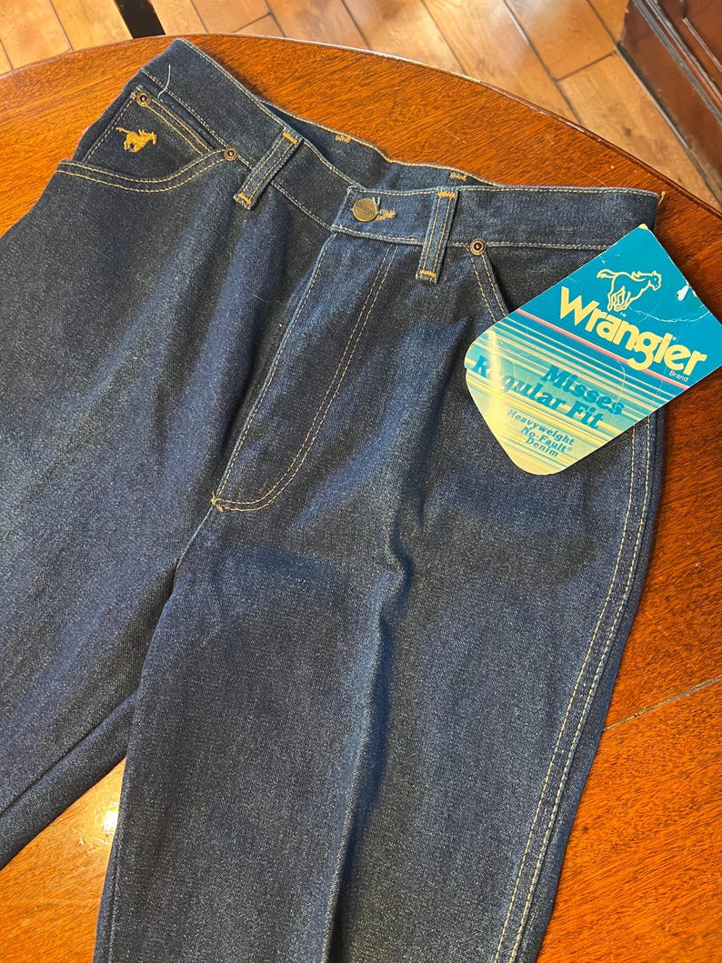 May include: Dark blue denim Wrangler jeans with a gold embroidered horse logo on the pocket. The jeans have a classic five-pocket design, a button and zipper closure, and a "Wrangler" brand tag. The jeans are labeled "Misses Regular Fit Heavyweight No-Fault Denim."