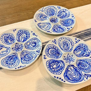 May include: Three white ceramic oyster plates featuring hand-painted blue designs. Each round plate has six recessed sections for individual oysters and a central circular compartment decorated with a blue floral pattern and blue shell motifs.