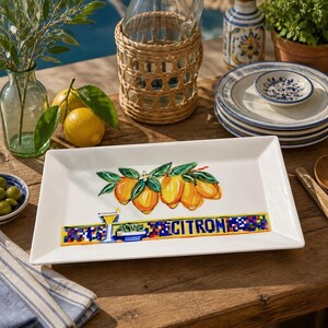 May include: A rectangular white ceramic serving platter with a vibrant lemon design and the word "CITRON" in colorful mosaic lettering. The platter is surrounded by other tableware and lemons, suggesting a summer theme.