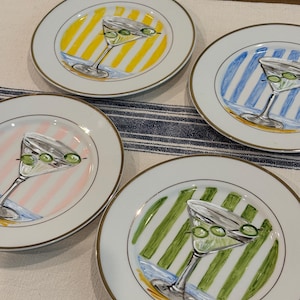 May include: A set of four white ceramic plates with a metallic silver rim. Each plate features a central illustration of a martini glass containing green olives, set against a background of vertical stripes in yellow, blue, pink, or green.
