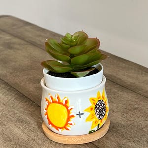 May include: A white ceramic planter with a sunflower and sun design. The planter has a wooden base and is filled with a green succulent plant.