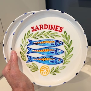 May include: A white ceramic plate featuring a hand-painted design of three blue sardines stacked horizontally, surrounded by a green leafy wreath. The word SARDINES is written in red above the fish, with a yellow lemon slice below.