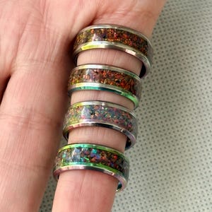 May include: Four rings with iridescent inlays are displayed on a finger. The rings have a silver band and a colorful, speckled inlay with hues of red, green, and purple. The rings are a unique jewelry item.