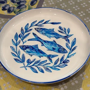 May include: White ceramic plate with a blue rim, featuring three stylized blue fish surrounded by blue leaf designs. The plate is set against a yellow and green patterned tablecloth, creating a coastal or nautical theme.