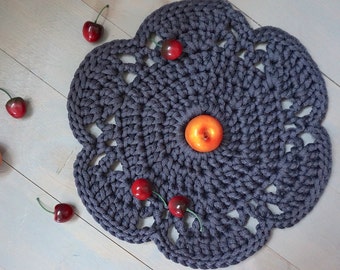May include: A dark gray crocheted doily with a circular design and intricate details. The doily is decorated with red cherries and a single orange apple.