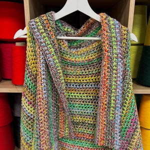 Op de afbeelding: Een veelkleurige gehaakte vest met een capuchon, hangend aan een witte hanger. De vest is gemaakt met verschillende kleuren, waaronder groen, blauw, geel en rood.