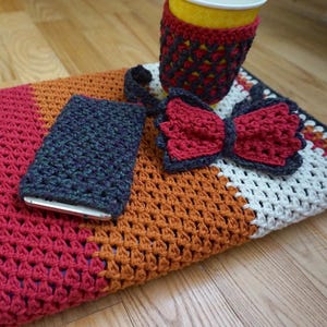 Könnte beinhalten: Eine mehrfarbige gehäkelte Decke mit einem Handy-Etui, einem Becherwärmer und einer Fliege. Die Decke besteht aus verschiedenen farbigen Quadraten, darunter Rot, Orange, Weiß und Creme. Das Handy-Etui ist dunkelgrün und schwarz, der Becherwärmer ist rot und schwarz und die Fliege ist rot.