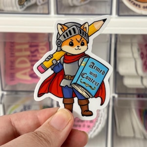 May include: A sticker featuring a cartoon fox in knight's armor, holding a pencil and a book titled "Armed with Context." The fox wears a helmet, red cape, and blue pants. The sticker is held in front of a clear storage container.