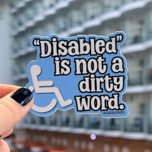 May include: A light blue sticker with the text "Disabled is not a dirty word." The sticker features a white wheelchair symbol. The background is blurred, showing a building with multiple balconies.