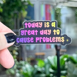 May include: A sticker with the text "today is a great day to cause problems" in a gradient of colors, held by a hand with a black painted nail. The sticker has a black border and a cloud-like shape.