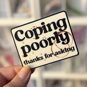 May include: A rectangular sticker with the words "Coping poorly" in a bold, black font, with coffee ring stains. The phrase "thanks for asking" is below. The sticker is held in a hand against a blurred background.