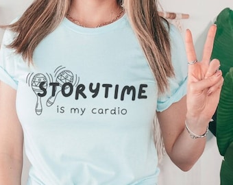 May include: Light blue t-shirt with the text "Storytime is my cardio" and a maraca graphic. The shirt is paired with blue jeans. The person is wearing a black and white polka dot headband.