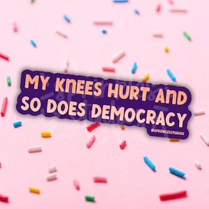 May include: A purple sticker with the text "MY KNEES HURT AND SO DOES DEMOCRACY" in white and orange. The sticker is surrounded by colorful sprinkles on a pink background. The sticker also includes the text "@SPRINKLESSTUDIOS".