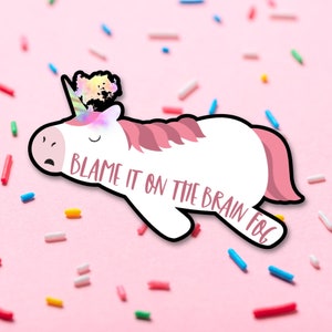 Op de afbeelding: Een witte eenhoornsticker met roze manen en staart, een regenbooghoorn en de tekst "Blame it on the Brain Fog". De sticker is zwart omlijnd en geplaatst tegen een roze achtergrond met kleurrijke hagelslag.