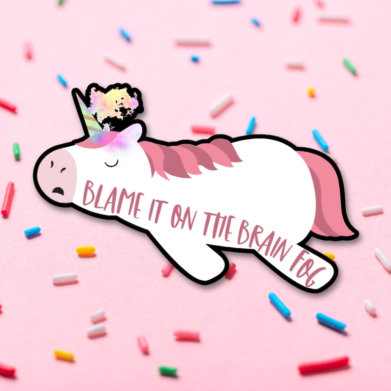 Op de afbeelding: Een witte eenhoornsticker met roze manen en staart, een regenbooghoorn en de tekst "Blame it on the Brain Fog". De sticker is zwart omlijnd en geplaatst tegen een roze achtergrond met kleurrijke hagelslag.