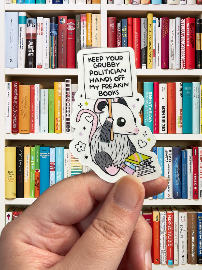 May include: A sticker featuring a cartoon possum holding a stack of books and a sign that reads "Keep your Grubby Politician Hands Off My Freakin Books".
